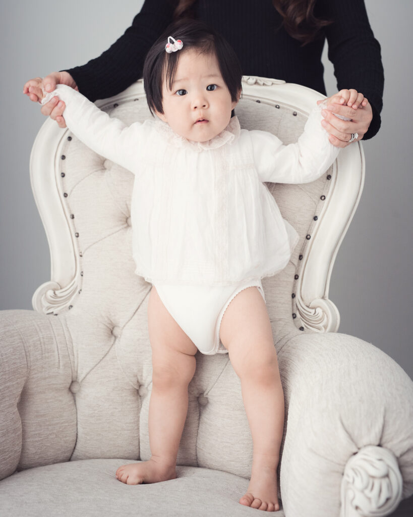 older baby standing on large chair hands held by unseeded adult standing behind chair