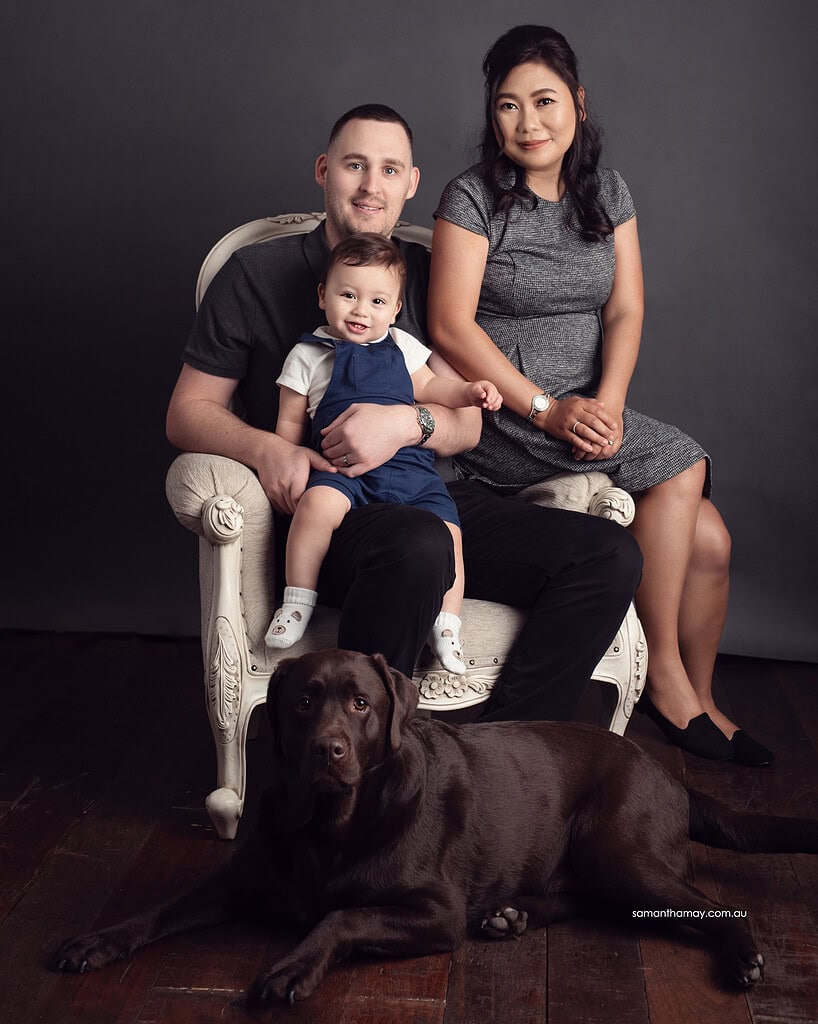 mother, father and toddler seated smiling to camera, chocolate colour family dog laying in front looking to camera